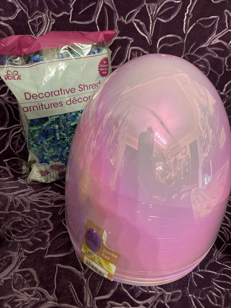 May include: A large iridescent pink plastic egg with a label that reads "Jumbo Egg". The egg is sitting on a purple fabric surface.