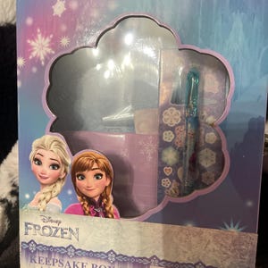May include: A Disney Frozen Keepsake Box Set. The box features Elsa and Anna from the movie. The set includes a sticker sheet, gel pen, notecards, envelopes, lock, and keys. The box is blue, purple, and white.