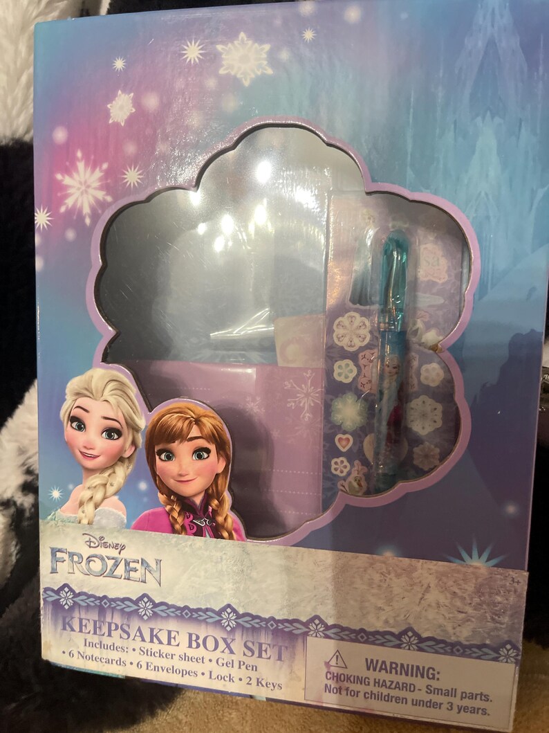 May include: A Disney Frozen Keepsake Box Set. The box features Elsa and Anna from the movie. The set includes a sticker sheet, gel pen, notecards, envelopes, lock, and keys. The box is blue, purple, and white.