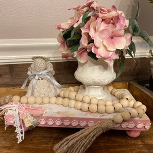 May include: A pink and white tiered tray with a wooden base, decorated with a white crocheted angel figurine, a wooden bead garland, and a pink floral print. The tray is accented with a burlap tassel and lace trim.