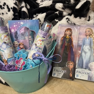 May include: A light blue metal basket filled with Frozen-themed gifts, including two dolls, markers, and a notebook. The basket is tied with a purple ribbon and sits on a black and white patterned blanket.