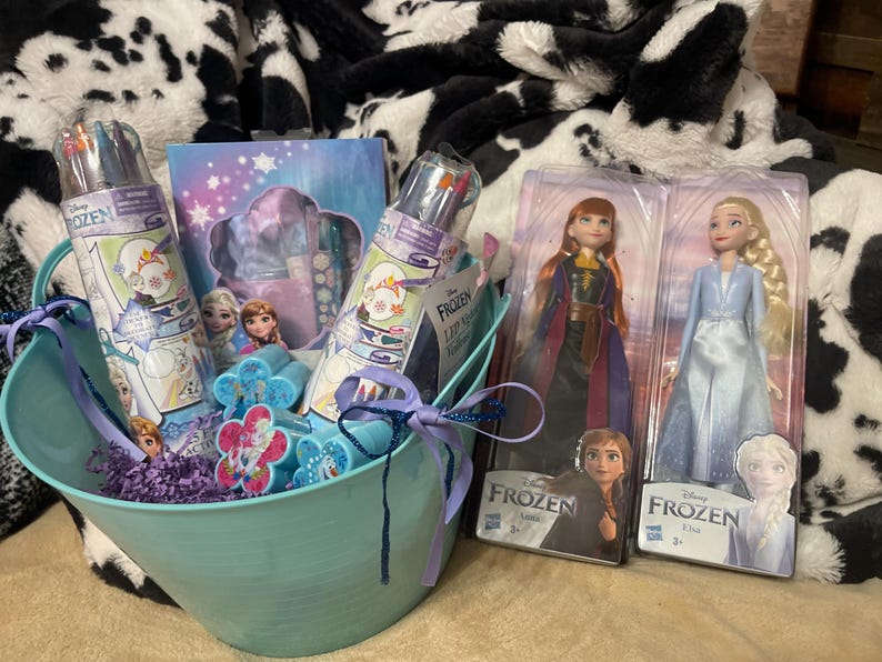 May include: A light blue metal basket filled with Frozen-themed gifts, including two dolls, markers, and a notebook. The basket is tied with a purple ribbon and sits on a black and white patterned blanket.