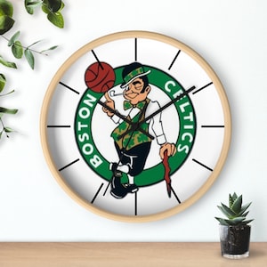 May include: A white clock with a black wooden frame and black hands. The clock face features a green circle with the Boston Celtics logo, which includes a cartoon leprechaun holding a basketball and a stick. The text "BOSTON" and "CELTICS" are written around the circle.