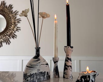 May include: A collection of home decor items featuring a black and white marbled pattern. This set includes a flat-bottomed vase with wooden flower reeds, two pillar candle holders with lit taper candles, and a small, low-profile candle holder.