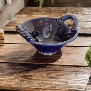 May include: A handmade, blue ceramic bowl with a glossy finish. The bowl features a unique, twisted handle design on each side. The interior has a dark, mottled pattern with hints of red and green. The bowl sits on a rustic wooden surface.
