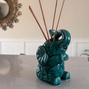 May include: A teal ceramic elephant-shaped diffuser with brown reeds. The elephant has detailed carvings and a glossy finish. The diffuser is on a light-colored surface, with a sunburst mirror in the background.