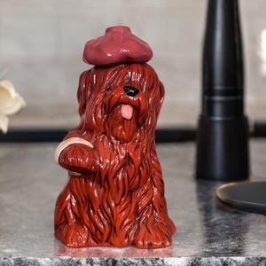 May include: A ceramic figurine of a dog with long, red fur, wearing a red beret and holding a white object. The dog has a pink tongue sticking out. The figurine is on a gray surface.
