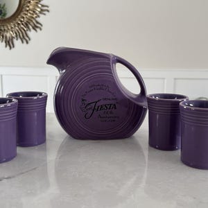 May include: A purple Fiesta pitcher with a handle and four matching tumblers. The pitcher has a circular design and the Fiesta logo. The tumblers are cylindrical with horizontal ridges. The set is on a white surface.