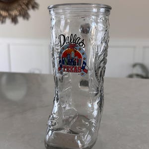 May include: Clear glass boot-shaped shot glass with embossed floral designs. The glass features a colorful Dallas, Texas, emblem. The shot glass is designed to resemble a cowboy boot.