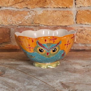 May include: A decorative ceramic bowl with an owl design. The bowl features a scalloped rim and a vibrant color palette of orange, blue, green, and red. The owl's face is centered on the bowl, with intricate details. The bowl is likely used for serving or display.