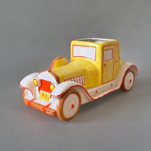 May include: A yellow ceramic car planter with white and red details. The car has a white windshield and a yellow door. The car has a red stripe on the front bumper and the number "1930" on the front.