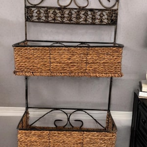 May include: A two-tiered wall-mounted storage shelf. The frame is dark metal with decorative scrollwork at the top and bottom. Two woven baskets made of natural-colored fibers provide storage space.