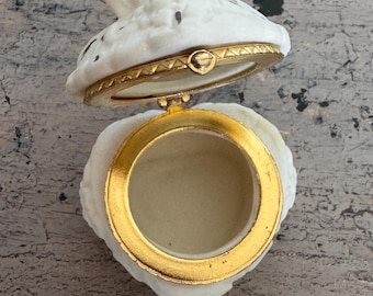 May include: A white, heart-shaped trinket box with a rose-shaped lid. The box has gold-colored trim and a hinged opening. The interior is a light beige color. The box is open, revealing the inside.