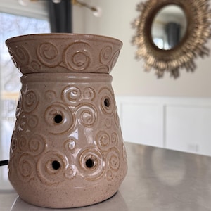 May include: A beige ceramic essential oil warmer with a two-piece design. The top piece is a bowl, and the bottom has several holes and a decorative swirl pattern. The warmer sits on a white countertop.