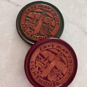 May include: Two circular tokens featuring a relief design of the Cape May Point Lighthouse. The top token has a dark green border with a copper-colored center, while the bottom token has a deep red border with a matching copper-colored center.