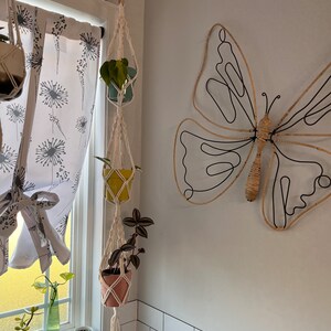 May include: Three hanging plant pots with macrame plant hangers, a pink, a yellow, and a blue pot, with plants in them, hanging from a pipe on a white tiled wall with a black metal butterfly wall decor.