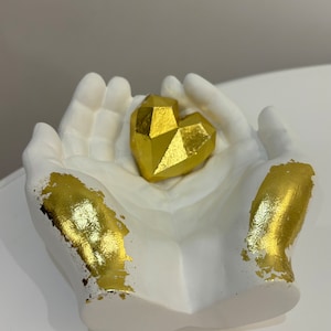 以下が含まれることがあります： 金色のアクセントが付いた白いセラミックの手が、金色の幾何学的なハートの彫刻を持っています。