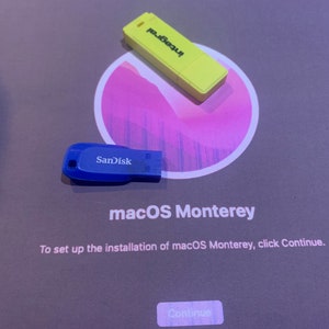 May include: A close-up of a computer screen showing the macOS Monterey installation screen. The screen is gray with white text. The text says "To set up the installation of macOS Monterey, click Continue." There is a blue SanDisk USB drive and a yellow USB drive on the screen.