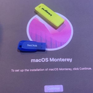 Könnte beinhalten: Nahaufnahme eines Computerbildschirms, auf dem der Installationsbildschirm von macOS Monterey angezeigt wird. Der Bildschirm ist grau mit weißem Text. Der Text lautet "Um die Installation von macOS Monterey einzurichten, klicken Sie auf Weiter". Auf dem Bildschirm befinden sich ein blauer SanDisk-USB-Stick und ein gelber USB-Stick.