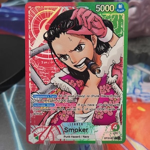 Op de afbeelding: Een ruilkaart met een karakterillustratie in roze en wit. De kaart heeft de tekst "Smoker" en "Punk Hazard / Navy". De kaart heeft een groene en rode rand, met het nummer "5000" rechtsboven. Voor een kaartspel.