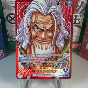May include: A red trading card depicting Silvers Rayleigh from One Piece. The card shows a portrait of Rayleigh with white hair and a beard. Text includes "5000", "LEADER", and game instructions. The card is displayed on a clear stand.