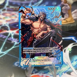 May include: A blue One Piece trading card featuring the character Rocks D. Xebec with a power level of 5000 and 5 life points. The card displays the text When Attacking You may trash 1 card from your hand Reveal 1 card from the top of your deck.