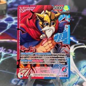 May include: A trading card featuring a character with a white beard, gold helmet, and red cape. The card has a 5000 power rating and includes game text. The card is labeled "LEADER Lucy" and "Dressrosa/Revolutionary Army."