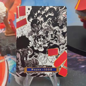 Puede incluir: Una tarjeta coleccionable con una ilustración en blanco y negro de personajes de anime, resaltada con detalles rojos. La tarjeta muestra el texto "DONK CARD" y "Power +1000" en una pancarta azul y dorada.