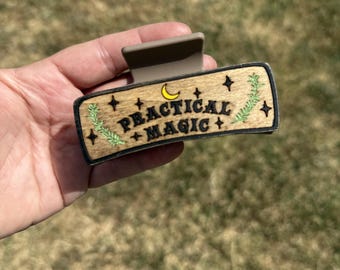 May include: A rectangular hair claw clip with a light wood-toned front panel featuring the black text "PRACTICAL MAGIC" centered between two green leafy sprigs, a small yellow crescent moon, and several black four-pointed star symbols.