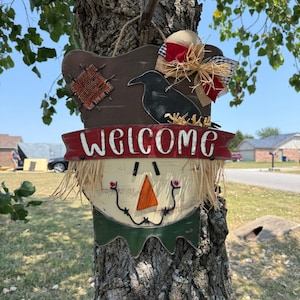 May include: A decorative scarecrow wall sign featuring a brown hat with a black crow silhouette, a red banner with the white text "WELCOME", a triangular orange nose, raffia hair, and a burlap bow with red and black plaid ribbon accents.
