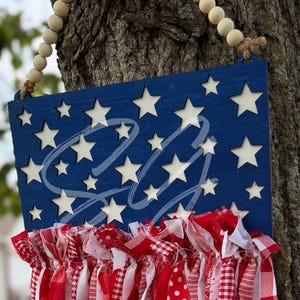 May include: A patriotic wooden sign with a blue background and white star cutouts. The sign has a red, white, and blue fabric fringe at the bottom and a beaded wooden hanger. The sign has the letters "LL" in a cursive font.