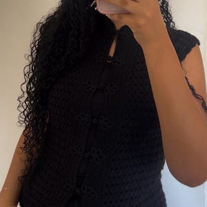 Puede incluir: Un top de ganchillo negro sin mangas con cuello alto tipo mandarín y cierre frontal con cinco botones decorativos de nudo. La prenda está confeccionada con un patrón de punto calado y texturizado en todo el cuerpo y el dobladillo.