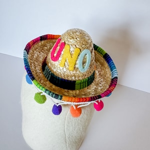 May include: A straw sombrero featuring a colorful woven fabric trim around the brim and crown. The word UNO is spelled out in felt letters in pink, yellow, and light blue on the front. A white string with hanging multi-colored pom-poms.
