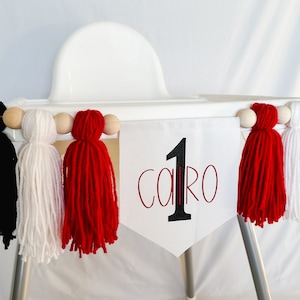 May include: A white high chair banner with the number "1" and the name "Cairo" in black and red lettering. The banner is decorated with black, white, and red yarn tassels and wooden beads.