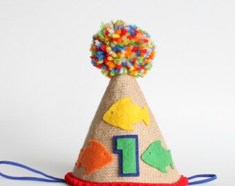 May include: A festive birthday party hat with a burlap cone, adorned with felt fish in yellow, orange, and green. The number "1" is displayed on the front. A colorful pom-pom tops the hat, and a blue string is attached.