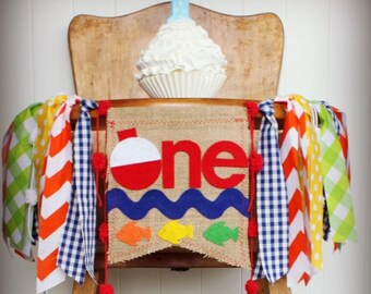 May include: A rustic high chair decorated for a first birthday. A burlap banner with a red and white bobber, the word "one", and fish. A cupcake with white frosting and a number one candle sits on top. Colorful fabric streamers hang from the chair.