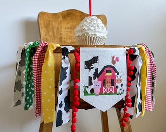 May include: A wooden high chair decorated with a farm-themed banner and colorful ribbons. The banner features a barn, farm animals, and the number one. A cupcake with white frosting and a red candle sits on the chair.