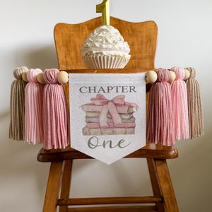 May include: A wooden high chair decorated for a first birthday. A cupcake with a gold number one candle sits on the chair. A banner with the words "Chapter One" and a stack of books is attached to the chair, along with pink and tan tassels.