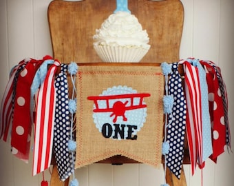 May include: A wooden chair decorated for a first birthday celebration. A cupcake with a blue number one candle sits on the chair. A burlap banner with a red aeroplane and the word "ONE" hangs below, with red, white, and blue ribbons.