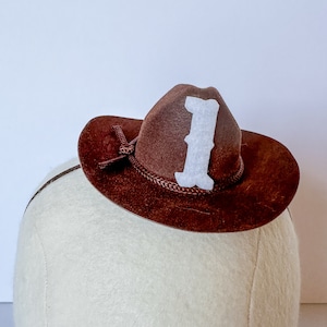 May include: A miniature brown cowboy hat with a white letter "I" on the front. The hat has a decorative rope band and a small brim. The hat is approximately 6 inches in diameter.