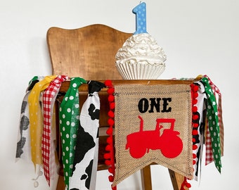 May include: A high chair decorated for a first birthday. A burlap banner with a red tractor and the word "ONE" is strung across the front. A cupcake with a blue number one candle sits on top. Colorful fabric streamers hang from the chair.