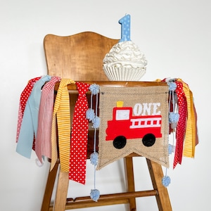 May include: A high chair decorated for a first birthday. A burlap banner with a red fire truck and the word "ONE" is draped across the front. A cupcake-shaped candle with a blue number one is on top.