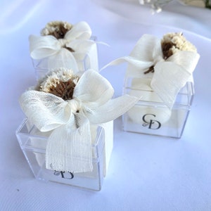 Op de afbeelding: Drie transparante acrylboxen met witte strikken en gedroogde witte bloemen. Elke doos bevat een wit rond voorwerp met de initialen "C & D" op de voorkant.