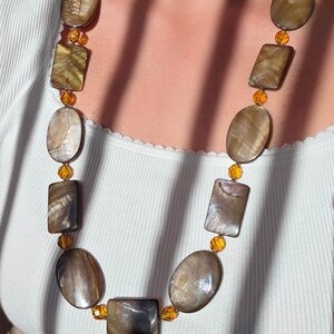 Op de afbeelding: Een lange ketting met grote, onregelmatig gevormde, iriserende bruine en grijze kralen. De kralen wisselen af tussen ovale en rechthoekige vormen, verbonden door kleine, amberkleurige kralen. De ketting wordt gedragen over een witte top.