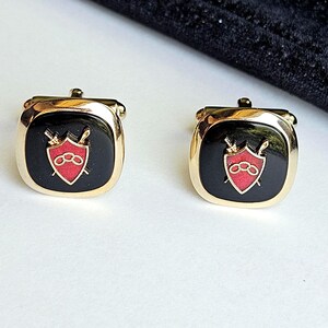 May include: A pair of gold-toned cufflinks with a black square face. Each cufflink features a red shield design with a sword and three rings. The cufflinks have a classic, elegant design.