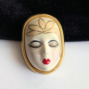May include: An oval-shaped brooch featuring a painted face with closed eyes, long eyelashes, and red lips. The face is framed by a gold-colored metal border. A delicate floral design adorns the forehead. The brooch has a vintage aesthetic.
