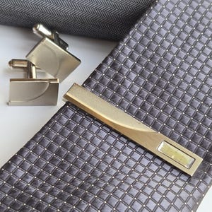 Op de afbeelding: Een set herenaccessoires met een patroon stropdas, manchetknopen en een stropdasclip. De stropdas is donkergrijs met een ruitpatroon en kleine witte stippen. Zilver.