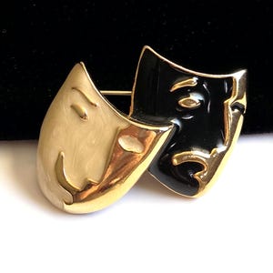 Peut inclure: Broche dorée représentant deux masques de théâtre stylisés. Un masque est crème, l'autre noir. Les deux masques ont des détails dorés et sont joints. La broche est probablement en métal et en émail.