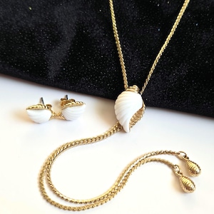 Könnte beinhalten: Ein goldfarbenes Halsketten- und Ohrring-Set. Die Halskette hat einen weißen Muschel-Anhänger und eine filigrane Kette. Die Ohrringe sind kleine Muschel-Ohrstecker. Das Schmuckset wird auf einer weißen Oberfläche mit schwarzem Hintergrund präsentiert.