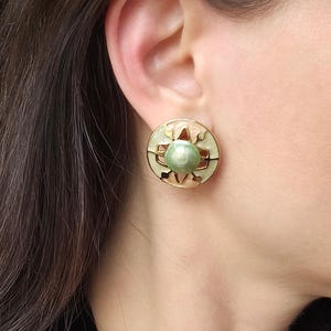 Könnte beinhalten: Ein Paar goldfarbene Ohrclips mit einem grünen und rosafarbenen Emaille-Design. Die Ohrringe haben eine runde Form mit einem zentralen grünen Perlen-Stein.